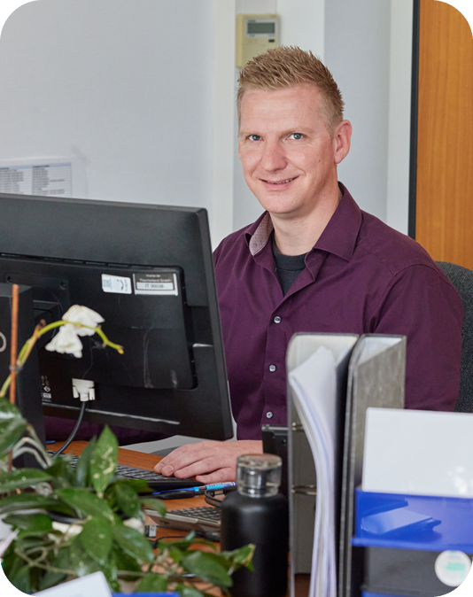 Teamleiter Kundenservice Freundlicher Mann mit auberginfarbenem Hemd sitzt am Computer und arbeitet